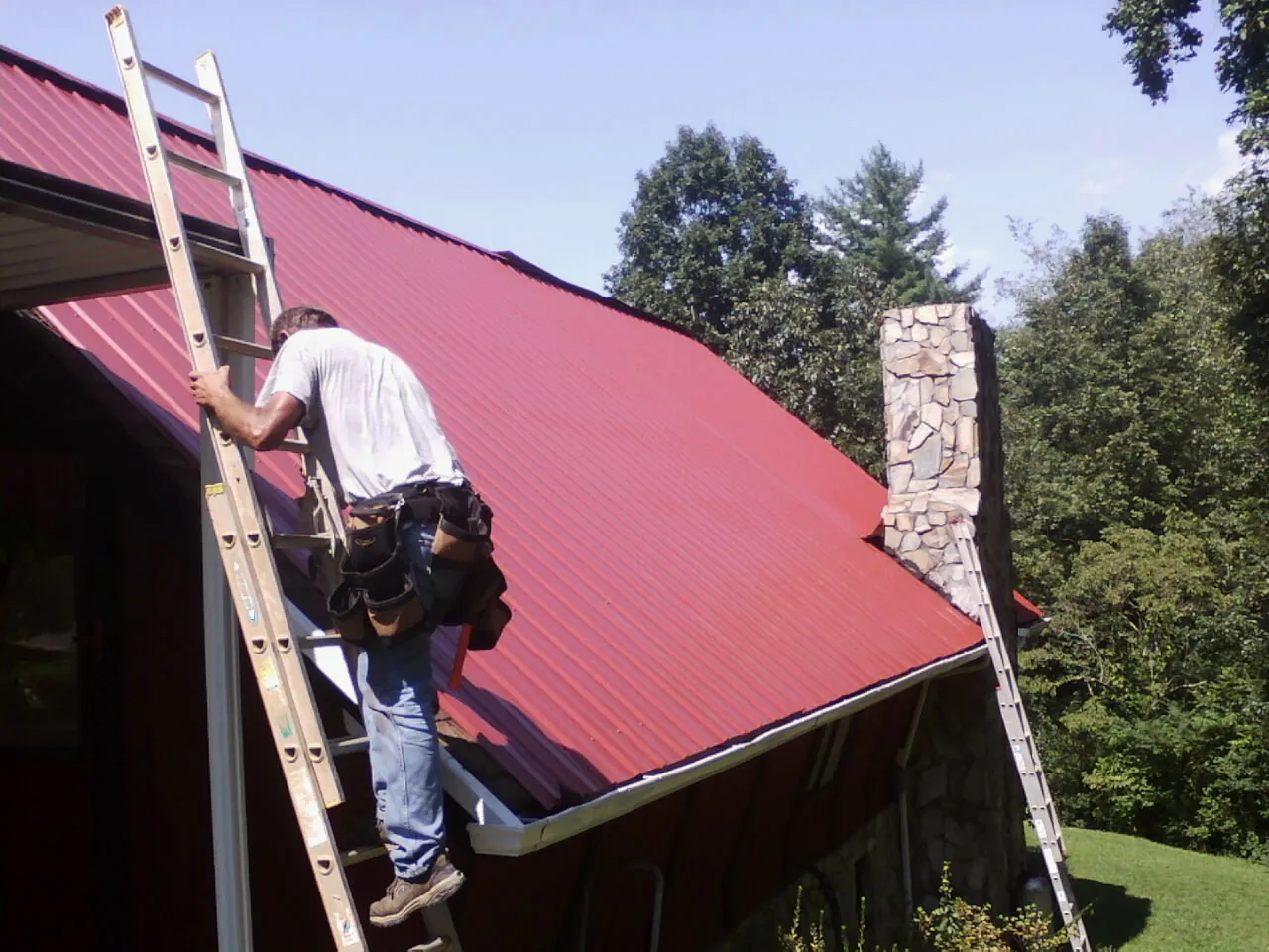 Roofer on a ladder beside a red metal roof and stone chimney — Soffit & Fascia Repair by Roofing Brothers in Harborcreek